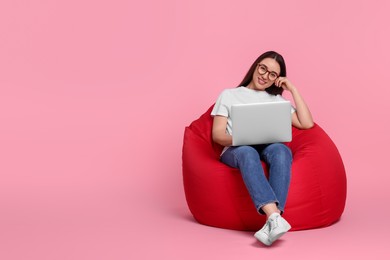 Photo of Smiling young woman with laptop sitting on beanbag chair against pink background, space for text