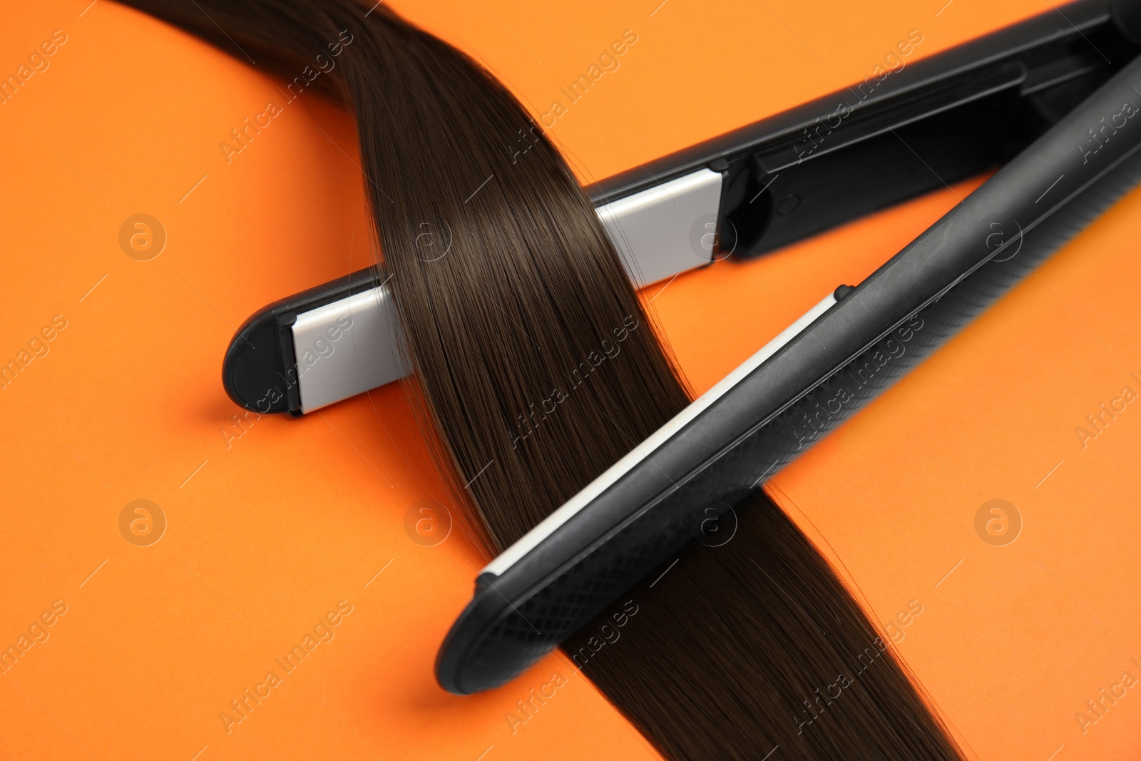 Photo of Straightener with brown hair lock on orange background, closeup