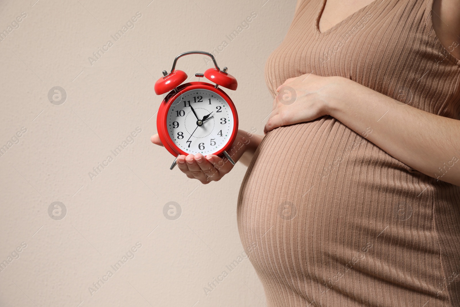 Photo of Young pregnant woman holding alarm clock near her belly on beige background, closeup and space for text. Time to give birth