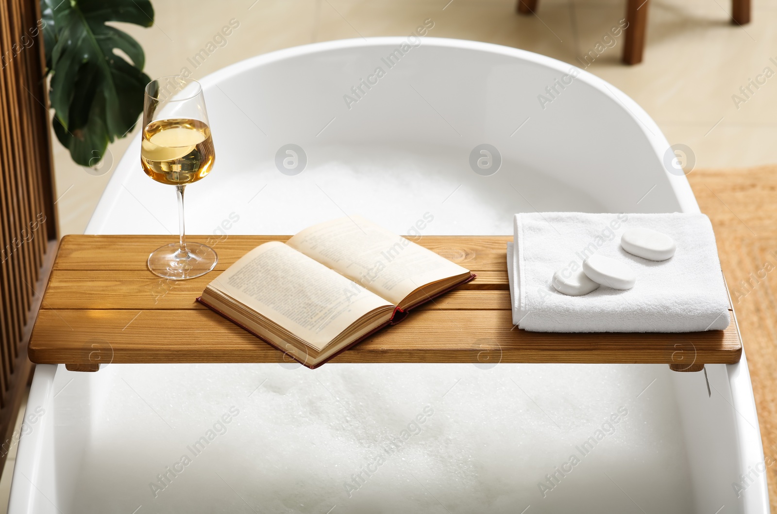 Photo of Wooden bath tray with glass of wine, open book, massage stones and towel on tub indoors. Relaxing atmosphere