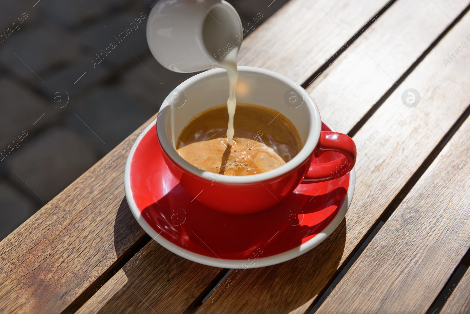 Photo of Pouring milk into cup of aromatic hot coffee at wooden table outdoors