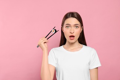 Photo of Emotional young woman holding sushi roll with chopsticks on pink background