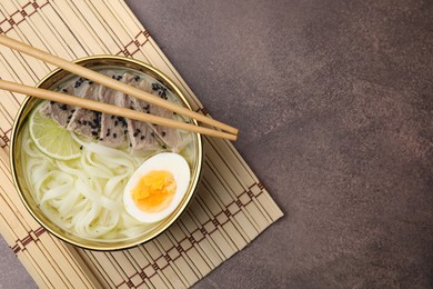 Photo of Bowl of delicious rice noodle soup with meat and egg on brown table, top view. Space for text