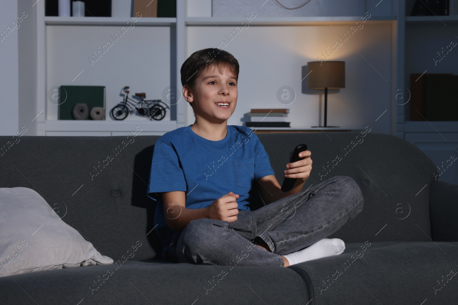 Photo of Emotional boy watching TV and holding remote control on sofa at home