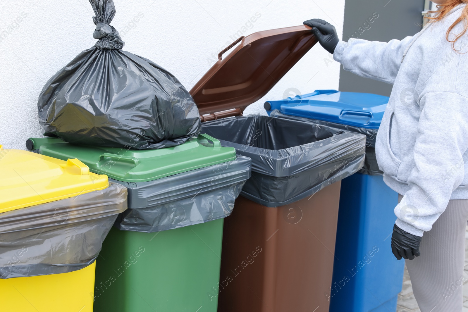 Photo of Woman opening bin to throw trash bag full of garbage outdoors, closeup. Recycling concept