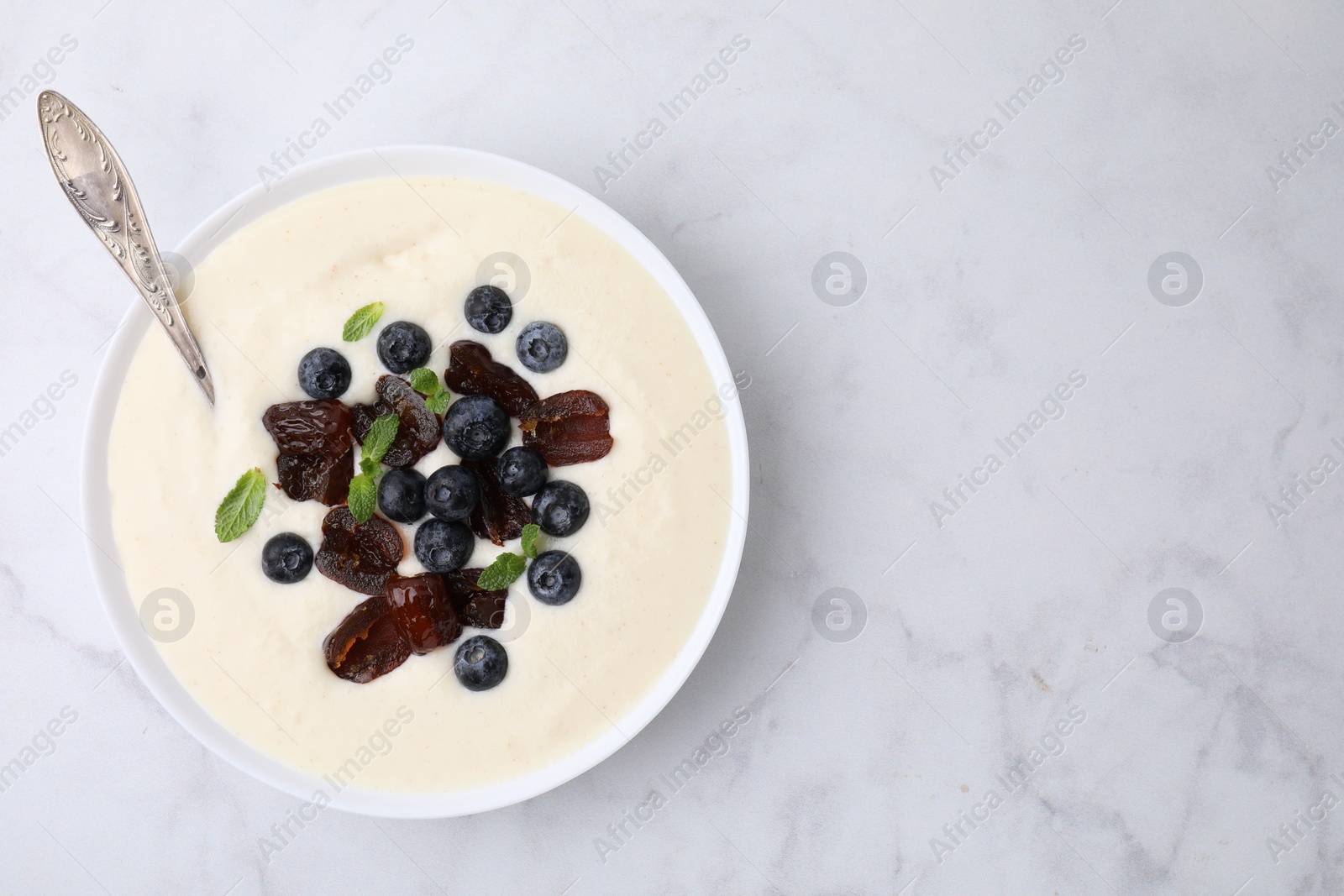 Photo of Delicious semolina pudding with blueberries, dates, mint and spoon in bowl on white marble table, top view. Space for text
