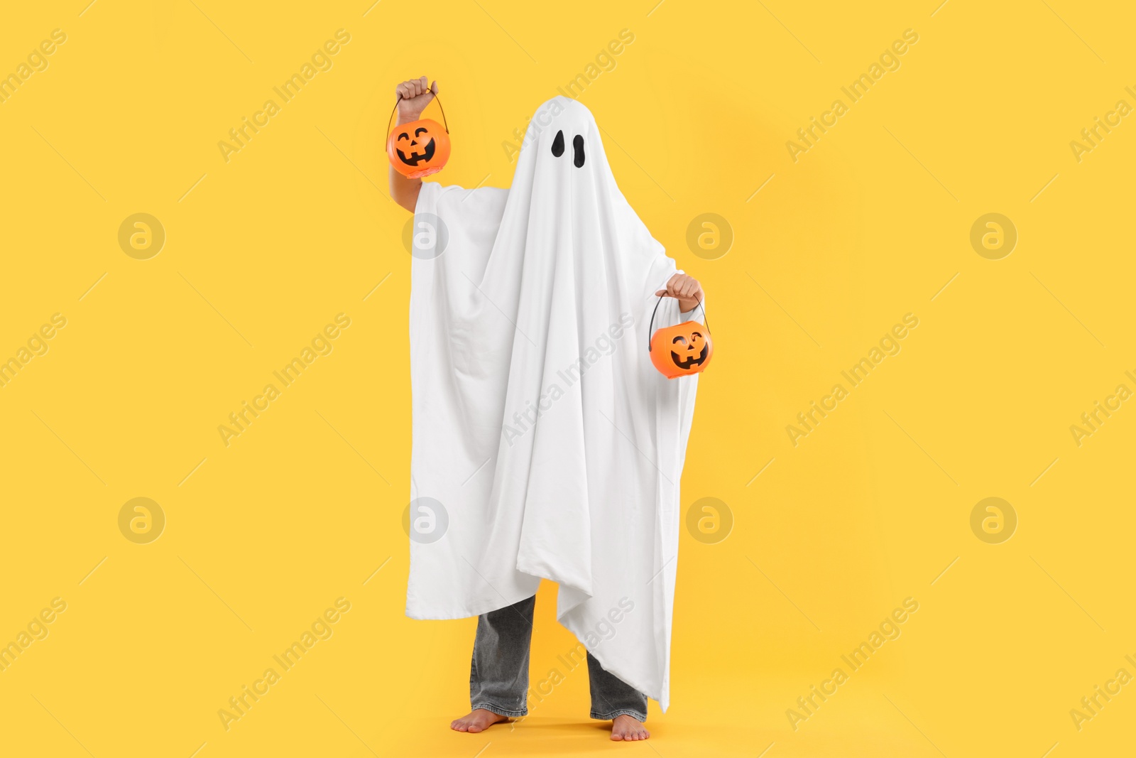 Photo of Woman in white ghost costume holding pumpkin buckets on yellow background. Halloween celebration