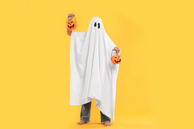 Photo of Woman in white ghost costume holding pumpkin buckets on yellow background. Halloween celebration