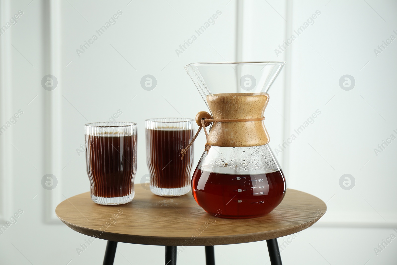 Photo of Glass chemex coffeemaker and glasses of coffee on wooden table against white wall