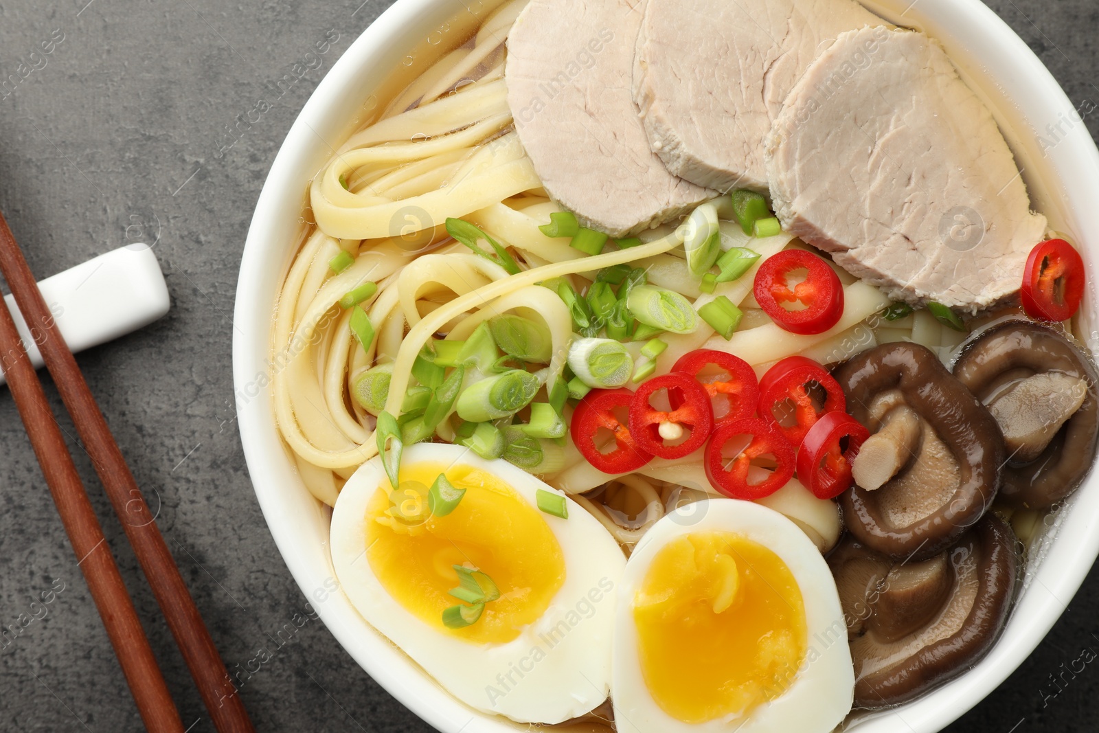 Photo of Delicious ramen with meat in bowl and chopsticks on grey textured table, top view. Noodle soup