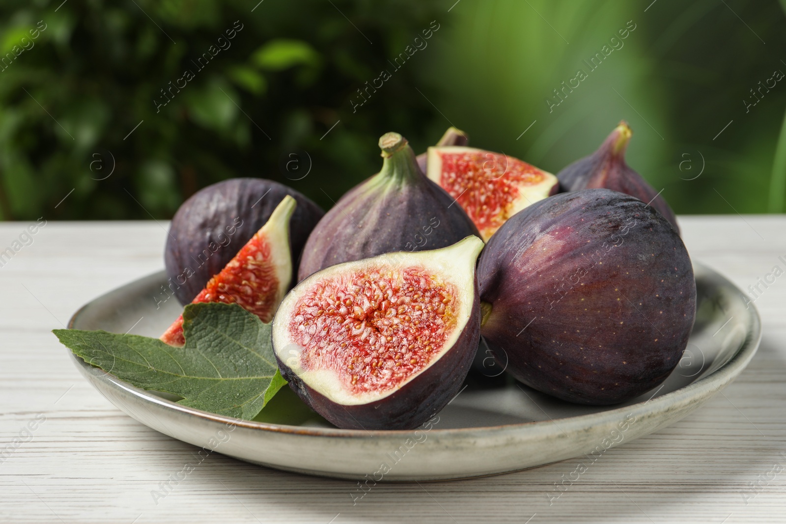Photo of Whole and cut ripe figs with leaf on white wooden table against blurred green background, closeup