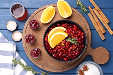 Photo of Cranberries in bowl, jars with sauce and ingredients on blue wooden table, flat lay