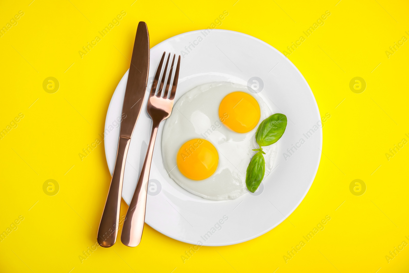 Photo of Tasty fried eggs with basil in plate on yellow background, top view