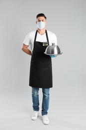 Photo of Waiter in medical face mask holding tray with lid on light grey background