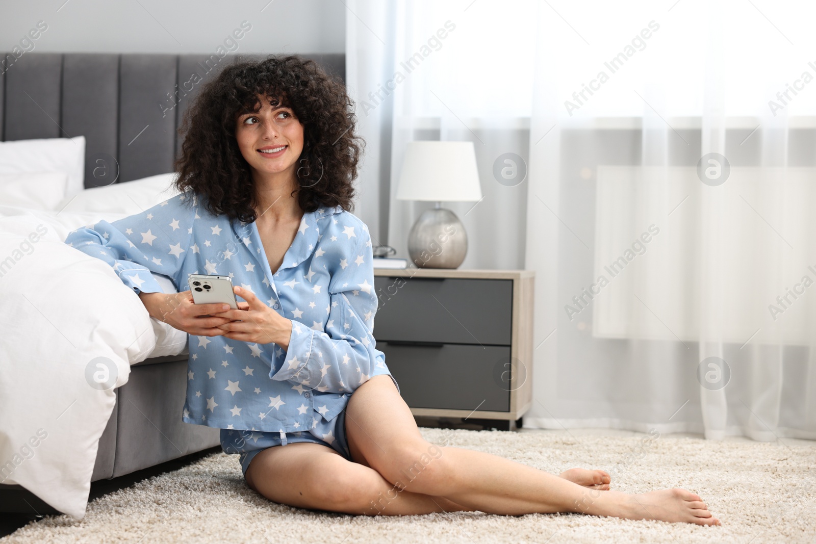 Photo of Beautiful young woman in stylish pyjama using smartphone on floor at home