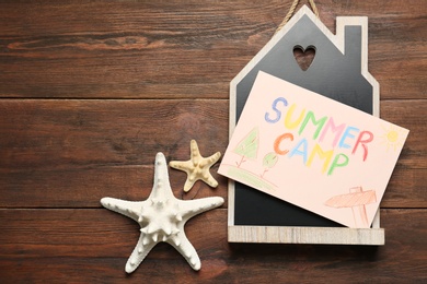 Card with text SUMMER CAMP, starfishes and house model on wooden table, flat lay