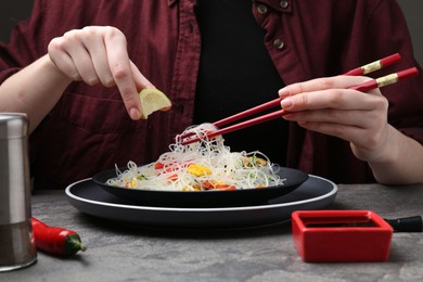 Photo of Stir-fry. Woman squeezing lime into tasty rice noodles with meat and vegetables at grey textured table, closeup