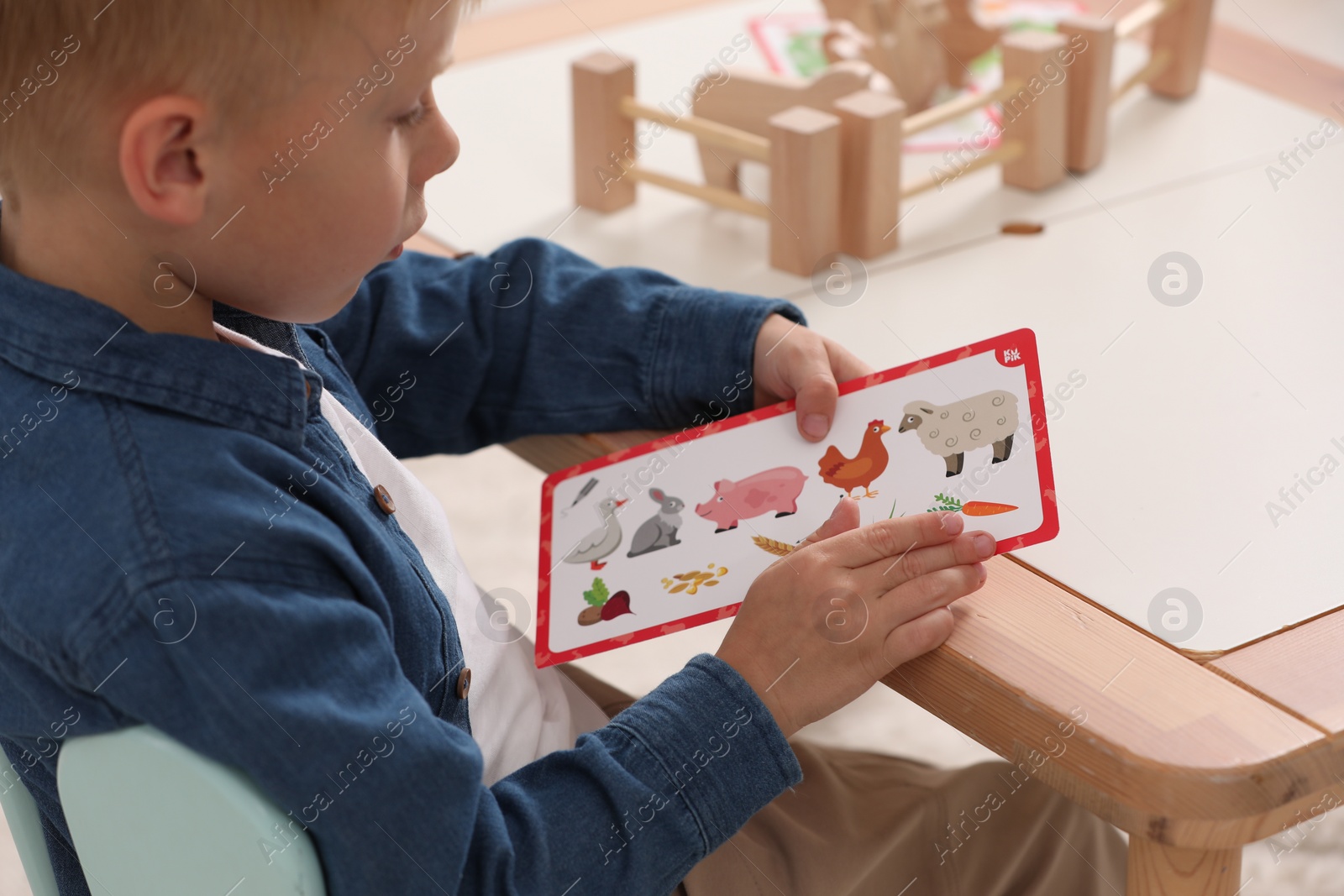 Photo of Little boy playing with set of wooden animals and fence at table indoors, closeup. Child's toy