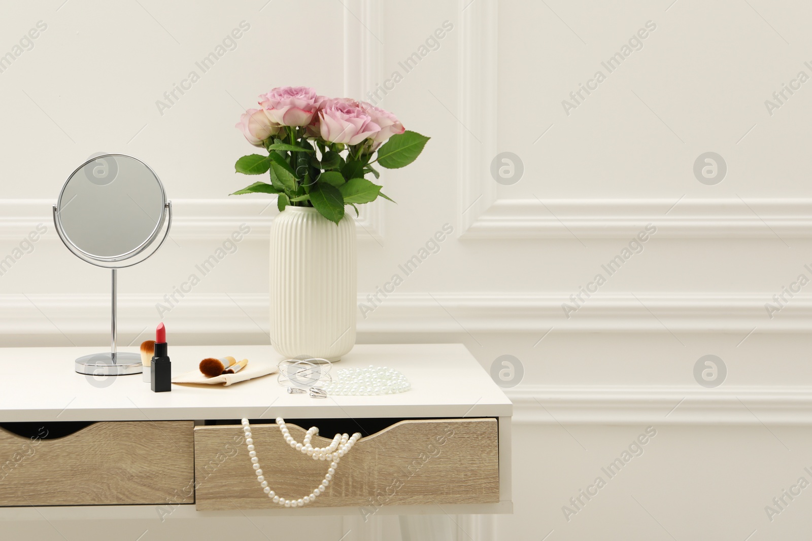 Photo of Mirror, cosmetic products, jewelry and vase with pink roses on white dressing table in makeup room, space for text
