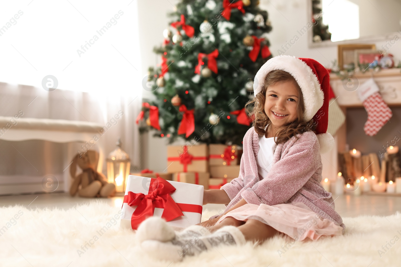 Photo of Cute little child in Santa hat with Christmas gift box at home