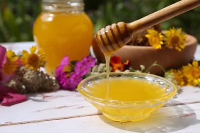 Delicious honey flowing down from dipper into bowl on white wooden table in garden, space for text