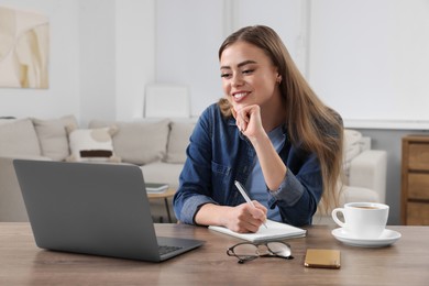 Photo of Happy woman with notebook using laptop at wooden table in room