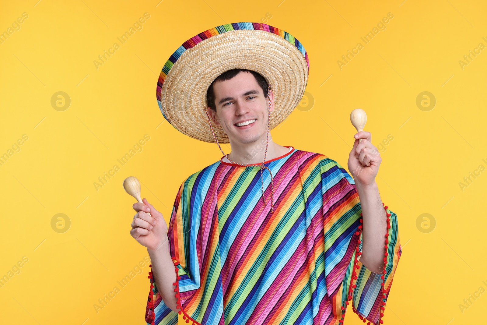 Photo of Young man in Mexican sombrero hat and poncho with maracas on yellow background