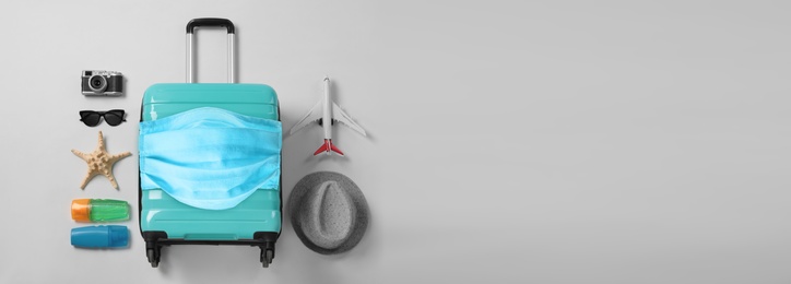 Image of COVID-19 pandemic, travel during coronavirus outbreak. Suitcase with protective mask and accessories on light grey background, flat lay 