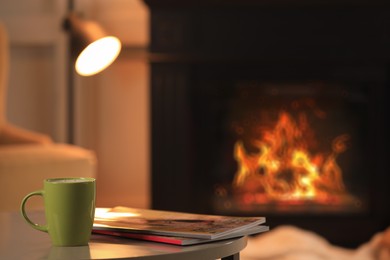 Photo of Cup of hot drink and magazines on table near fireplace at home, space for text. Cozy atmosphere