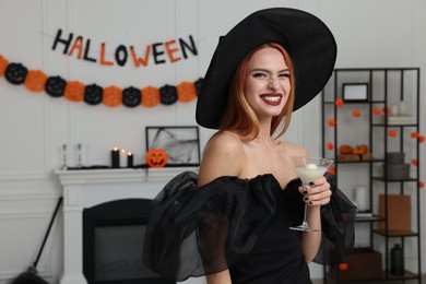 Photo of Happy young woman in scary witch costume with glass of cocktail indoors, space for text. Halloween celebration