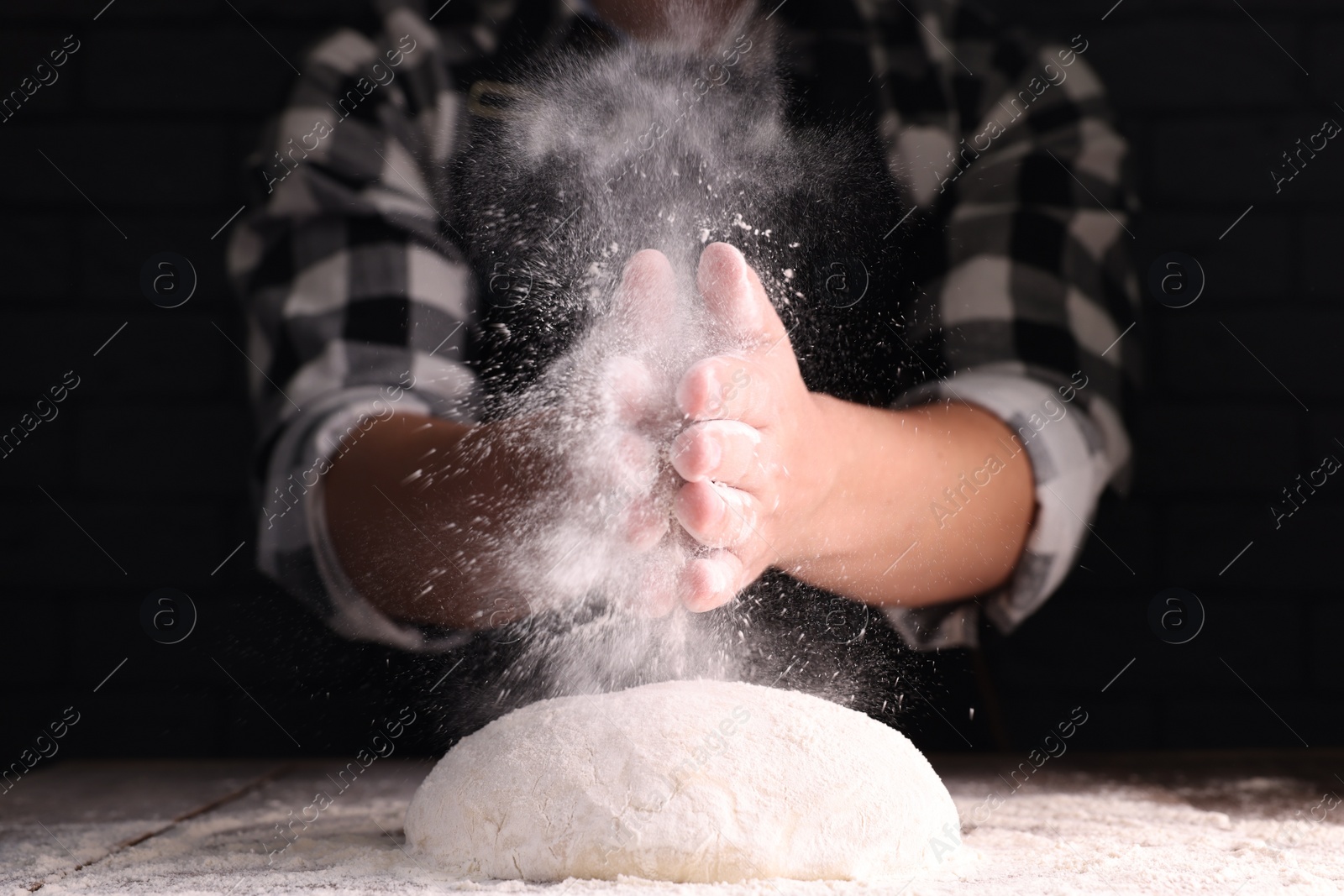 Photo of Man sprinkling flour over dough at wooden table on dark background, closeup