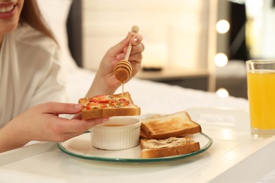 Young woman pouring honey onto toast near white tray on bed at home, closeup