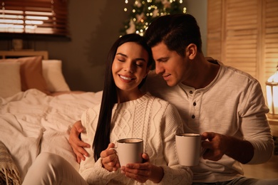Photo of Happy couple with cups in festively decorated bedroom. Christmas celebration