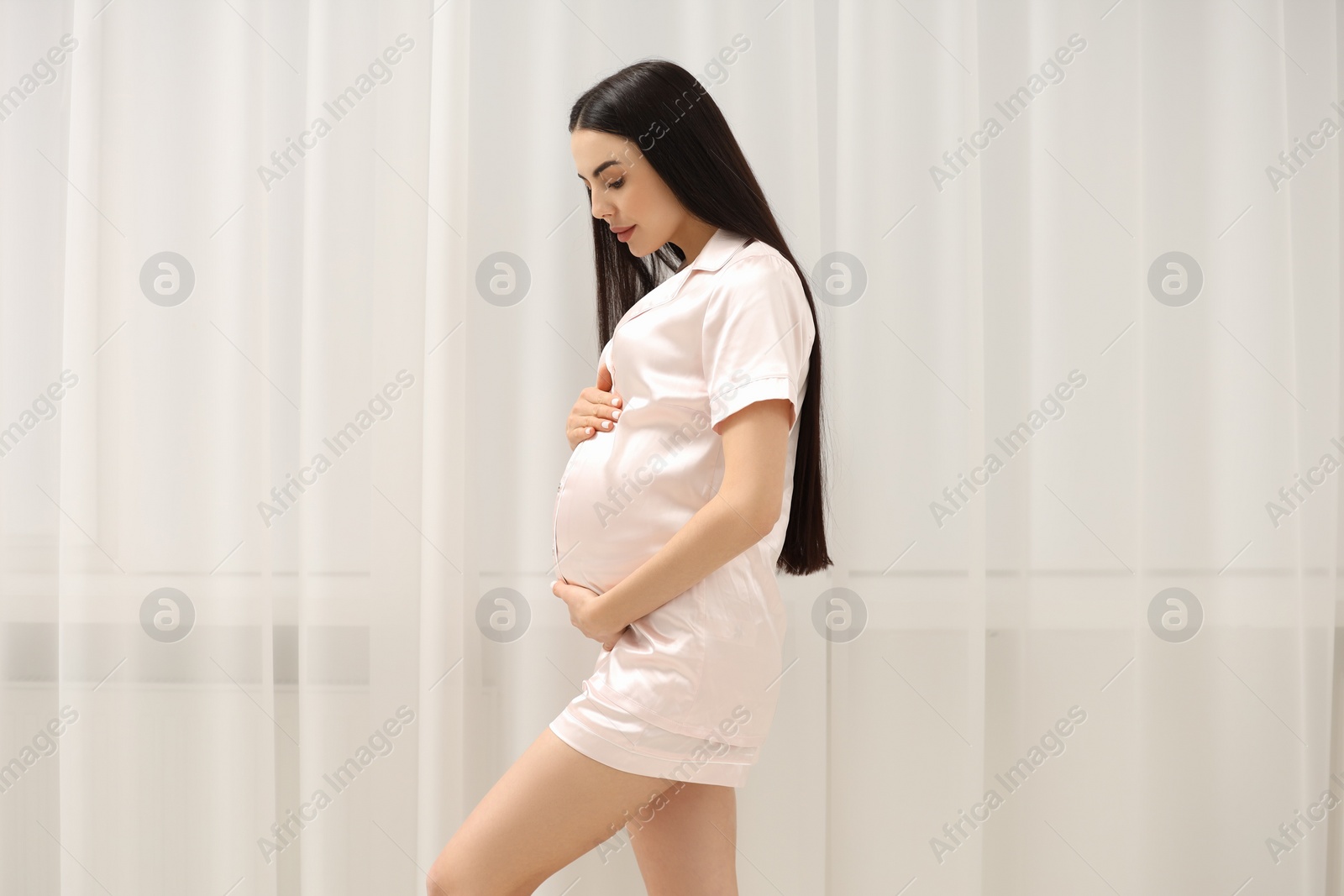 Photo of Pregnant young woman in pajamas near window indoors