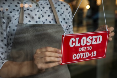 Photo of Woman in mask putting red sign with words Closed Due To Covid-19 hanging onto glass door, closeup