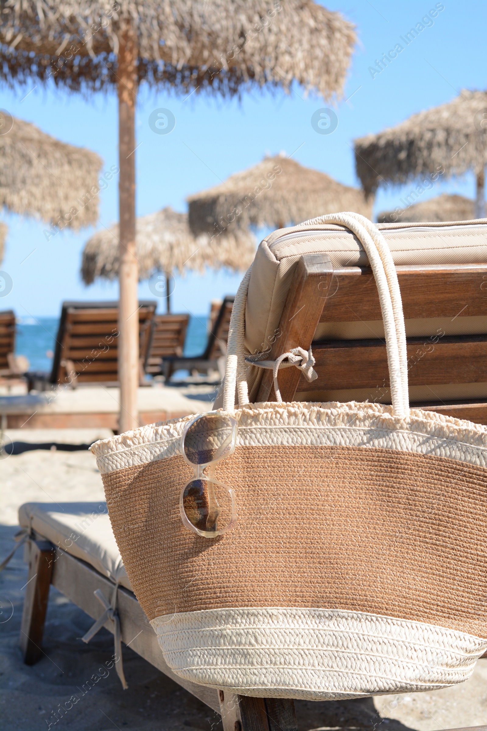 Photo of Straw bag with sunglasses on wooden sunbed near sea. Beach accessories