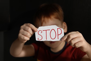 Photo of Little boy holding piecepaper with word Stop against black background, focus on hands. Domestic violence concept