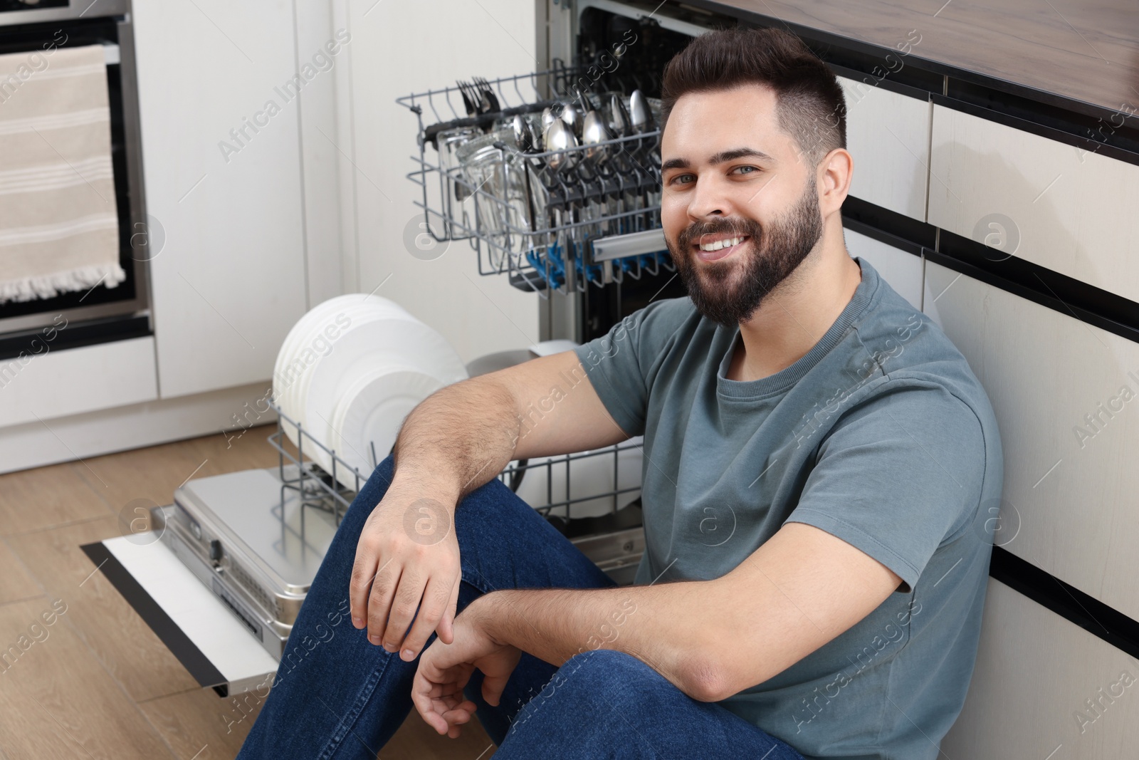 Photo of Smiling man sitting near open dishwasher in kitchen