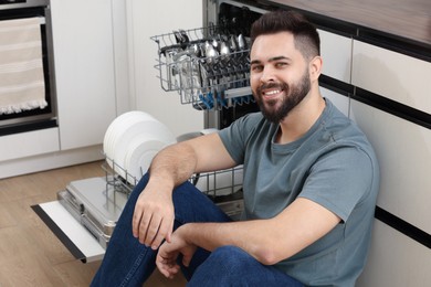 Smiling man sitting near open dishwasher in kitchen