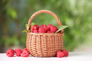 Wicker basket with tasty ripe raspberries and leaves on white table against blurred green background