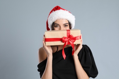 Woman in black dress and Santa hat holding Christmas gift on grey background