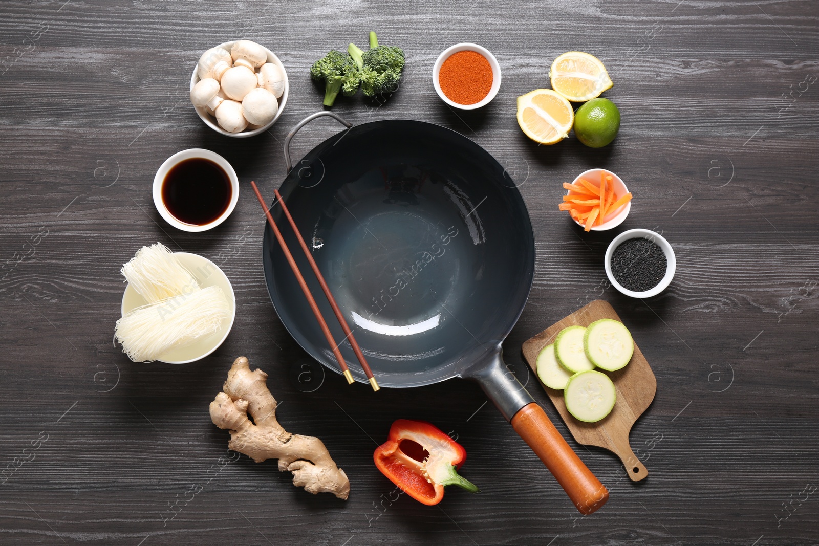 Photo of Empty iron wok and chopsticks surrounded by ingredients on dark grey wooden table, flat lay