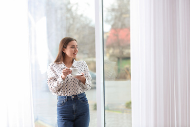 Photo of Young woman with cup near window indoors