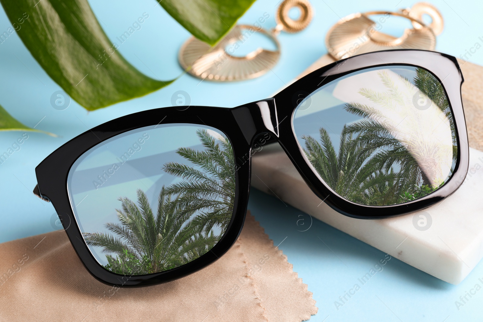 Image of Elegant sunglasses on light blue background, closeup. Sky and palm trees reflecting in lenses