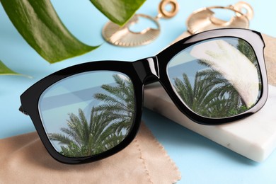 Image of Elegant sunglasses on light blue background, closeup. Sky and palm trees reflecting in lenses
