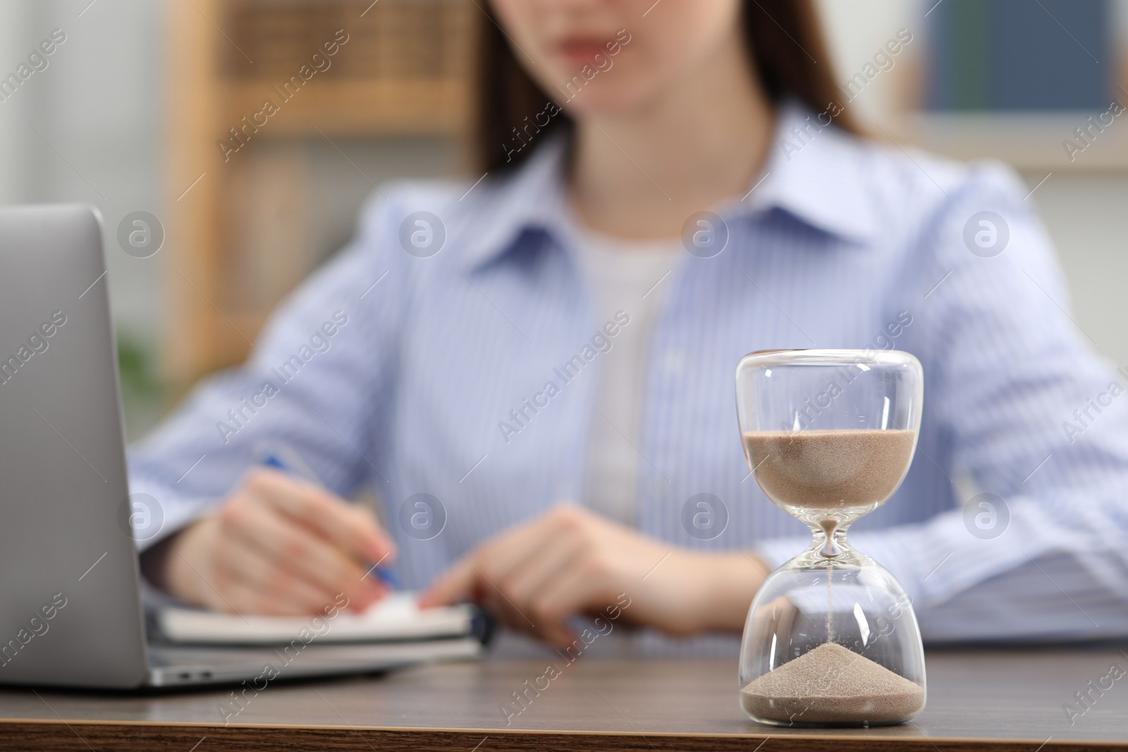 Photo of Hourglass with flowing sand on desk. Woman taking notes while using laptop indoors, selective focus