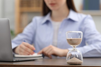 Photo of Hourglass with flowing sand on desk. Woman taking notes while using laptop indoors, selective focus