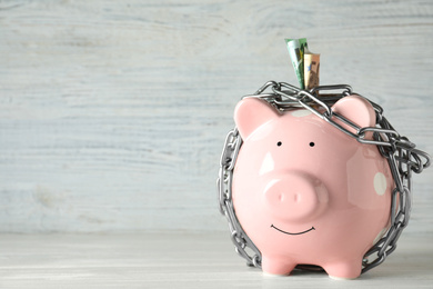 Photo of Piggy bank with steel chain and banknotes on white wooden table, space for text. Money safety concept