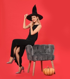 Photo of Beautiful woman wearing witch costume near armchair and pumpkin on red background. Halloween party