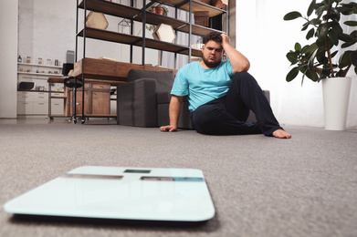 Photo of Scales and depressed overweight man on floor at home